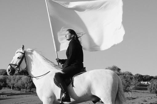 Das Schwarzweiß-Foto zeigt die Künstlerin Marina Abramović auf einem weißen Schimmel mit einer großen weißen Fahne in der Hand 