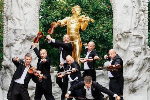 The photo shows the musicians of the Mnozil Brass ensemble in black tailcoats, violins in hand, posing in front of the golden Johann Strauss monument in Vienna's Stadtpark
