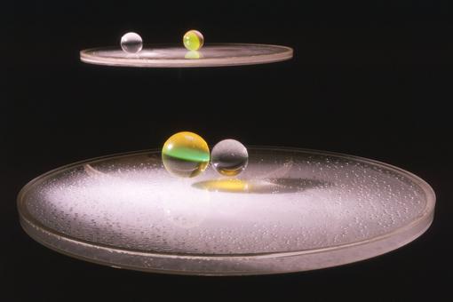 The photo shows an illuminated, pale pink turntable on which a glass plate lies, with two acrylic spheres floating above it, one of them yellow-green, all against a deep black background