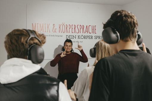 Photo of young people with headphones taken from behind. In front of the group is a young woman speaking in sign language