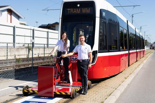 Zu sehen sind die beiden Teilnehmer:innen an der Tram-WM am 13.9.2025 in Wien: Elisabeth Urbanitsch und Florijan Isaku. Sie bfinden sich auf einer roten Draisine vor einer modernen Straßenbahn-Garnitur der Wiener Linien
