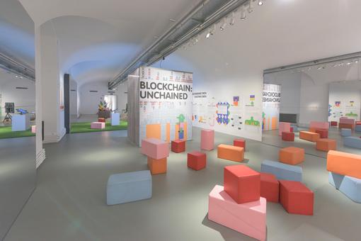 On display is part of the Blockchain:Unchained exhibition, the lettering can be read on a wall in the middle. The room contains large foam building blocks in different colors, with mirrors and texts on the walls to the left and right