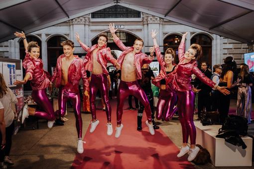 The photo shows a group of 3 women and 3 men in glittering pink costumes, all jumping in the air together and smiling at the camera