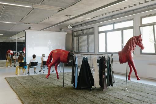 Das Foto zeigt eine künstlerischen Installation zum Angewandte Festival 2024. Zu sehen ist ein geteiltes, rotes Plastikpferd,  zwischen dem Kopf und dem Hinterteil des Pferdes befindet sich eine Kleiderstange mit Kleidungsstücken.