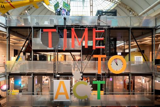 Ausstellungsansicht: über drei Ebenen des Museums ist der Schriftzug "Time to act" in großen Lettern zu lesen, daneben eine Uhr mit dem Zeiger auf fünf vor zwölf