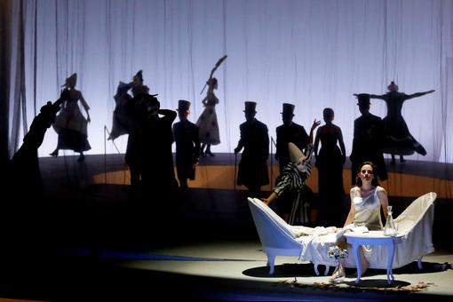 The scene photo from the opera “La traviata” by the Volksopre Vienna shows the leading actress Rebecca Nelsen in a white dress on a white chaise longue, in the background a crowd of people in the shade