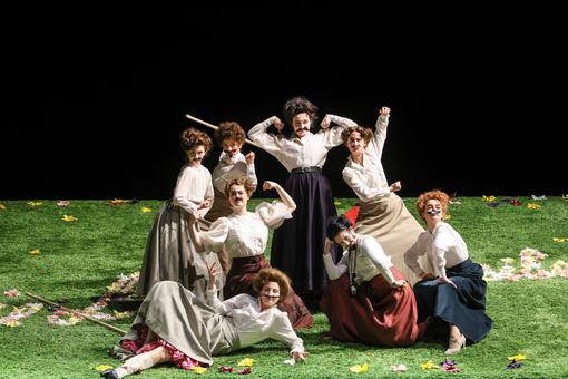 Scene photo from the opera "The Merry Wives of Windsor": Eight women in long skirts, presenting themselves in typical male poses and with glued-on moustaches
