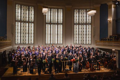 Das Foto zeigt die Wiener Bachsolisten und den BAchCHorWien auf der Bühne des Mozartsaales im Wiener Konzerthaus, dahinter bunte Jugendstil-Glasfenster