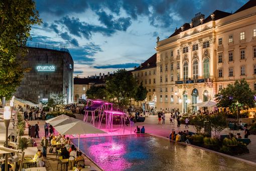 The photo shows the main courtyard of the MuseumsQuartier in an evening summer atmosphere, many people are in the main courtyard and in guest gardens, evening lighting