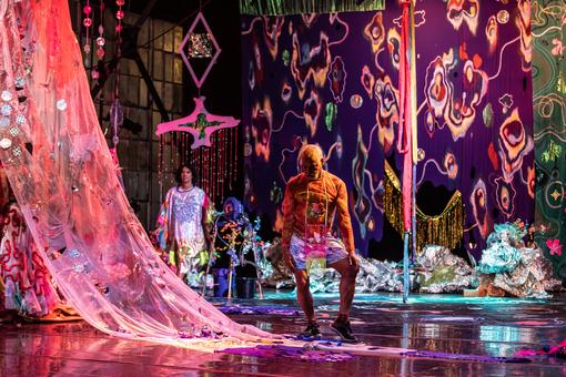 Das Szenebild zeigt eine Performance von TRY Collaborative / Circo Zero, auf einer sehr bunten, aufwendig dekorierten Bühnen, im Vordergrund ein Tänzer, der zu Boden blickt 