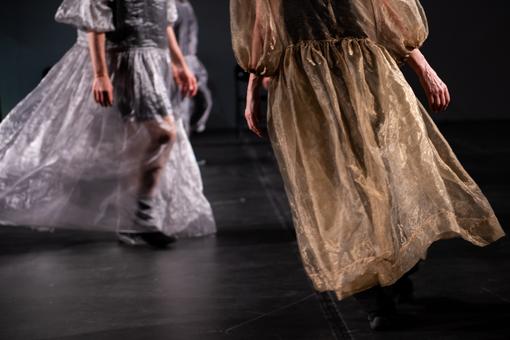 Das Szenefoto der Tanzperformance zeigt zwei Personen in bodenlangen Kostümen, die für Ballkleider stehen sollen, eines in Gold, das andere in durchscheinenden Silber