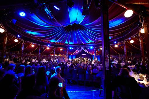 Das Foto zeigt eine Event-Location in blauer Beleuchtung: im Vordergrund das Publikum an eingedeckten Tischen, im Hintergrund ein Orchester auf einer Bühne