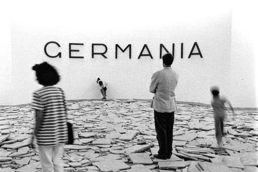 The black and white photo shows four people moving around on a floor covered with broken stone slabs and tiles. One man is looking at the black lettering Germania in capital letters on a white wall in the background