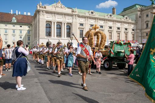 Das Foto zeigt den Einzug einer Blasmusikgruppe in Lederhosen und Tracht auf den Wiener Heldenplatz, auf einem Wagen im Zug befindet sich eine Erntedank-Krone aus Getreide