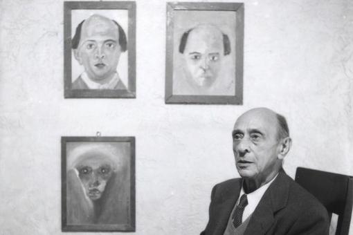 Das Schwarzweiß-Foto zeigt den Komponisten Arnold Schönberg sitzend, im Hintergrund an der Wand ist drei von ihm gemalte Bilder, davon zwei Selbstporträts, zu sehen