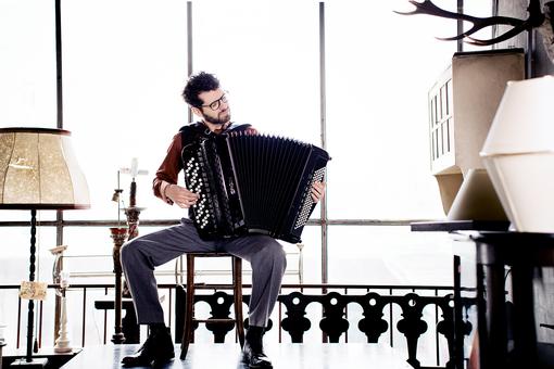 Photo of a man with a full beard and glasses sitting on an armchair and playing an accordion