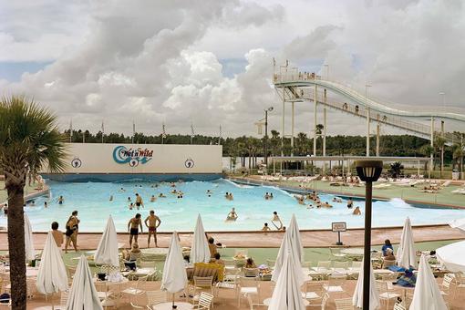 Farbfoto eines Aquaparks in Florida mit Swimmingpool, Wasserrutsche, im Vordergrund eine Terrasse mit weißen Gartenmöbeln und Sonnenschirmen