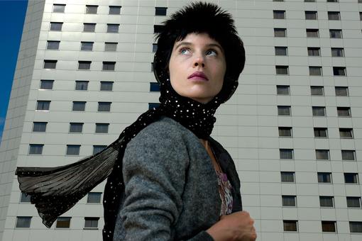 Photo of a woman in front of a light gray high-rise building with a monotonous facade. The woman is wearing a gray jacket, a black scarf and a black fur hat. She is looking upwards to the left.