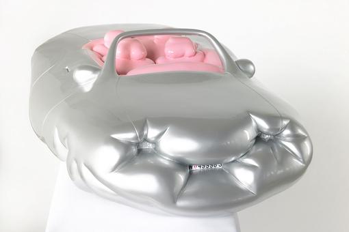 Das Foto zeigt eine Plastik, die aussieht wie ein aufblasbares, dickes Cabrio in der Farbe Silber, mit vier angedeuteten rosafarbenen Insassen
