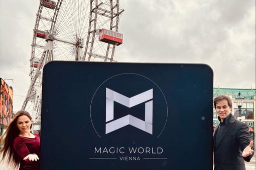 Das Foto zeigt die Magier Anca und Lucca, die ein Schild mit der Aufschrift Magic World Vienna und dem Logo der Location in die Kamera zeigen, im Hintergrund das Wiener Riesenrad