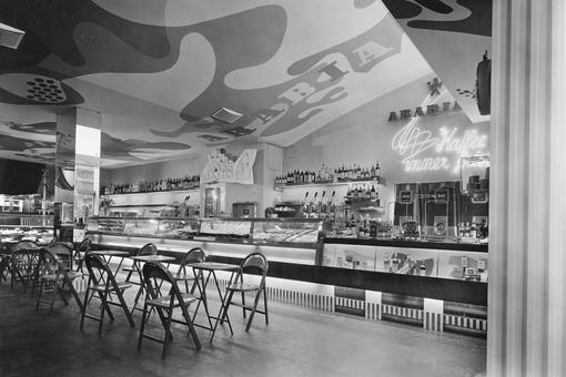 Black and white photo / Interior view of Café Arabia: a long sales counter and display cases, tables and armchairs in front of it, on the ceiling the lettering Arabia + design.