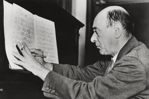 Black and white photo of Arnold Schoenberg sitting at a piano and writing notes in a patitur