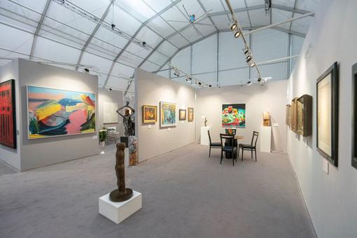 Exhibition view: Different paintings on light gray walls, in the foreground a small brown statue. The whole exhibition is located in a large tent
