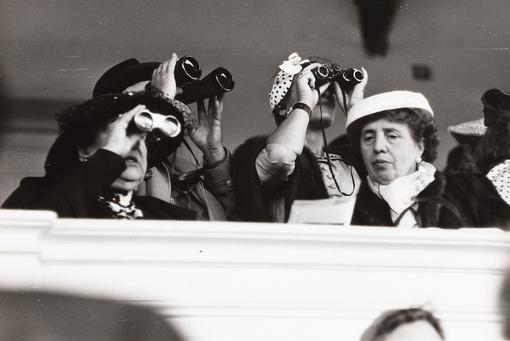 Black and white photo of three women at horse race all looking through binoculars