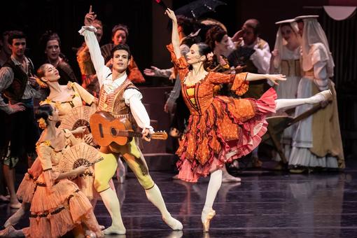 Tänzer:innen in spanischen Kostümen, im Vordergrund das Solistenpaar, die Ballerina in einem orangefarbenen Kostüm in Spitzen-Position, der Mann mit einer Gitarre in der Hand