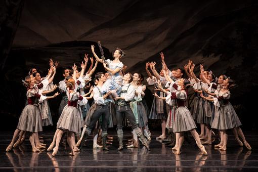 Group of dancers, the women wear Drindl costumes, the men wear lederhosen with white shirts, the ballerina in a light blue dirndl is carried on the shoulders of two men