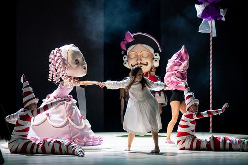 Das Foto zeigt die Hauptdarstellerin in einem weißem Kleid, die von einer übergroßen Puppe in rosa Kostüm an der Hand gehalten wird. Im Vordergrund zwei Tänzer als rotweiß gestreifte Würmer mit spitzen Hüten