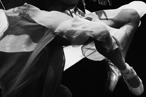 Schwarzweiß-Foto, das zwei gekreuzte Beine mit weißen Ballettschuhen zeigt, die von verschiedenen Lagen Tüll verdeckt werden