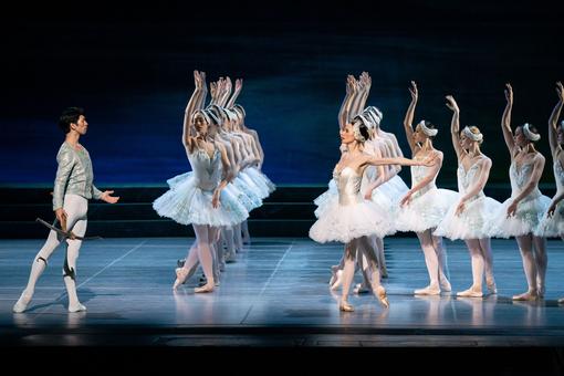 A formation of dancers in white costumes and white headdresses faces the solo dancer, who carries a crossbow