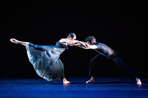 Photo of a ballet couple in blue costumes holding each other in position by the arms. The scene takes place on an intensely blue floor.