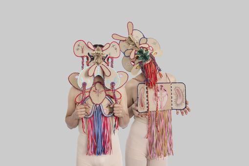 Das Foto zeigt zwei Personen in fleischfarbenen Trikots, die vor dem Kopf und dem Brustbereich aufwendig gestaltete Masken mit bunten Fransen tragen