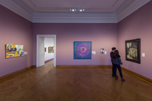 Exhibition room, pink-purple walls, white ceiling, herringbone parquet floor, 3 paintings on the walls, 2 visitors looking at a painting