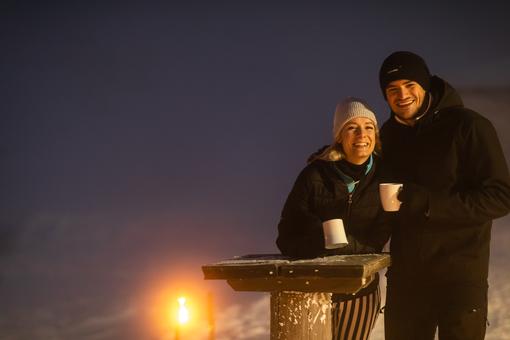 Das Foto zeigt ein Paar an einem hölzenen Stehtisch. Der Mann und die Frau tragen winterliche Kleidung halten jeweils ein weißes Häferl in Händen. Im Hintergrund leuchtet ein weihnachtliches Licht und taucht das Bild in eine besinnliche Stimmung