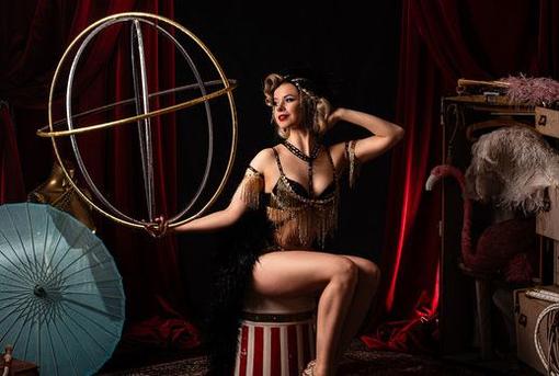 The photo shows a woman in a burlesque costume and 1920s hairstyle looking to the right at a globe indicated by metal circles, which she is holding in her hand.