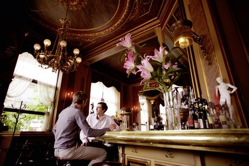Innenansicht des historischen und entsprechend eingerichteten Kaffeehauses, zwei Männer unterhalten sich am Tresen, im Vordergrund eine Vase mit üppigen rosafarbenen Lilien