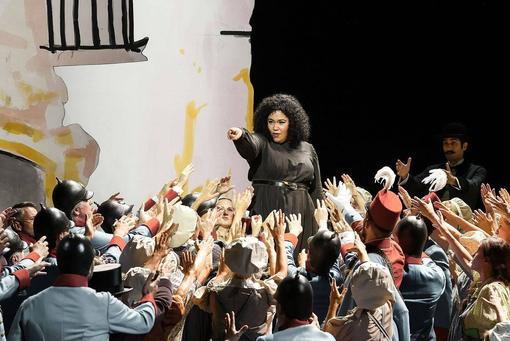 Scene photo from the opera Carmen: Carem stands in a black costume in an elevated position in a crowd of people, the people stretch their hands towards Carmen. Carmen points towards the camera with an outstretched index finger