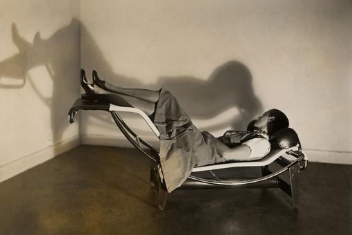 Schwarzweiß-Foto von der Designerin Charlotte Perriand, die auf einer Chaise longue, einem Design-Stück aus Metall mit Polsterung, liegt.