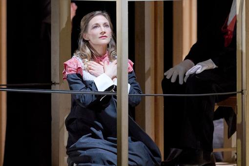 Das Szenefoto aus der Oper "Dialogues des Carmélites" zeigt eine der Sängerinnen in einem schwarzem Kleid mit rotrosa Kragen, die auf dem Boden kniet, mit den Händen auf der Brust überkreuzt