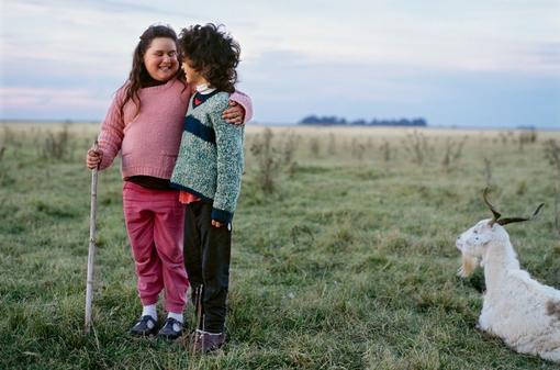 The photo shows two children in a spacious meadow with a white goat lying next to them. The girl smiles with her eyes closed, holds a stick in her hand and puts her left arm around the other child in a friendly manner