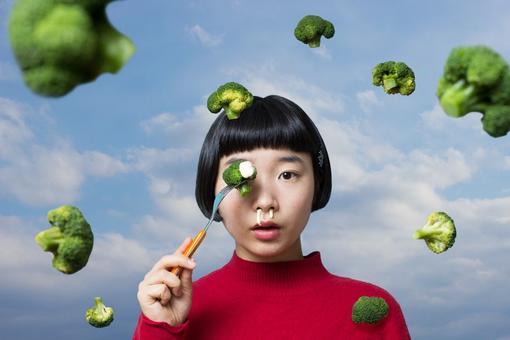 Photo of asian girl in red sweater holding fork with skewered broccoli in front of her left eye, white sauce running out of nose, broccoli flying around her