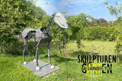 Foto einer Metallskulptur, die das Skelett eines Pferdes zeigt und in einem uppig grünen Garten steht