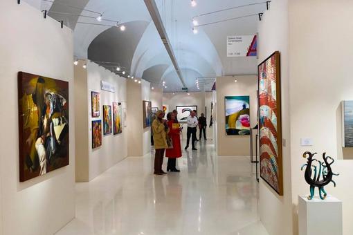 Exhibition view: Different paintings on light beige walls with small spotlights. Visitors:inside stroll through the exhibition