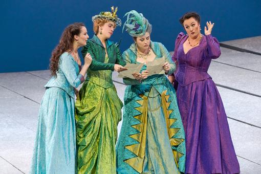 Das Szenefoto aus der Oper "Falstaff" zeigt vier der Sänger:innen in vier aufwendigen Kostümen in den Farben Grün, Hellblau, Türkis und Violett, die gemeinsam zwei Schriftstücke betrachten