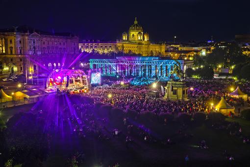 Photo with illuminated stage on which the Vienna Symphony Orchestra is playing, in the foreground the audience