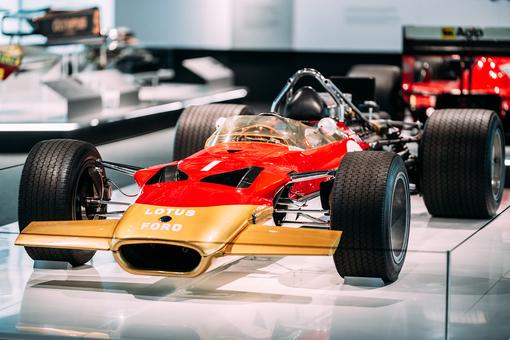 The photo shows a historic Formula 1® racing car in the colors red and gold standing on a shiny pedestal