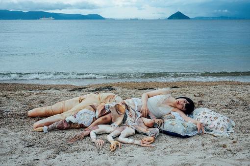 Photo of the Japanese artist, who is like an installation with arms and legs made of different materials spread around her, lying on pads on a beach, with the sea in the background.